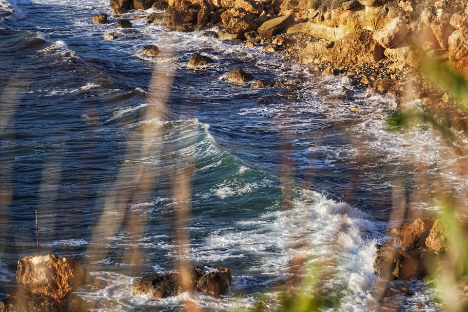 A photo of some waves crashing against rocks that can be seen in the top left of the image. Very out-of-focus grass stalks can be seen in the foreground.