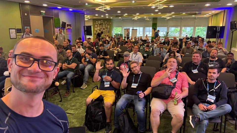 A selfie of myself smiling, positioned to the left of the image. Behind me is a room full of people sat down facing me. Some people at the front are waving.