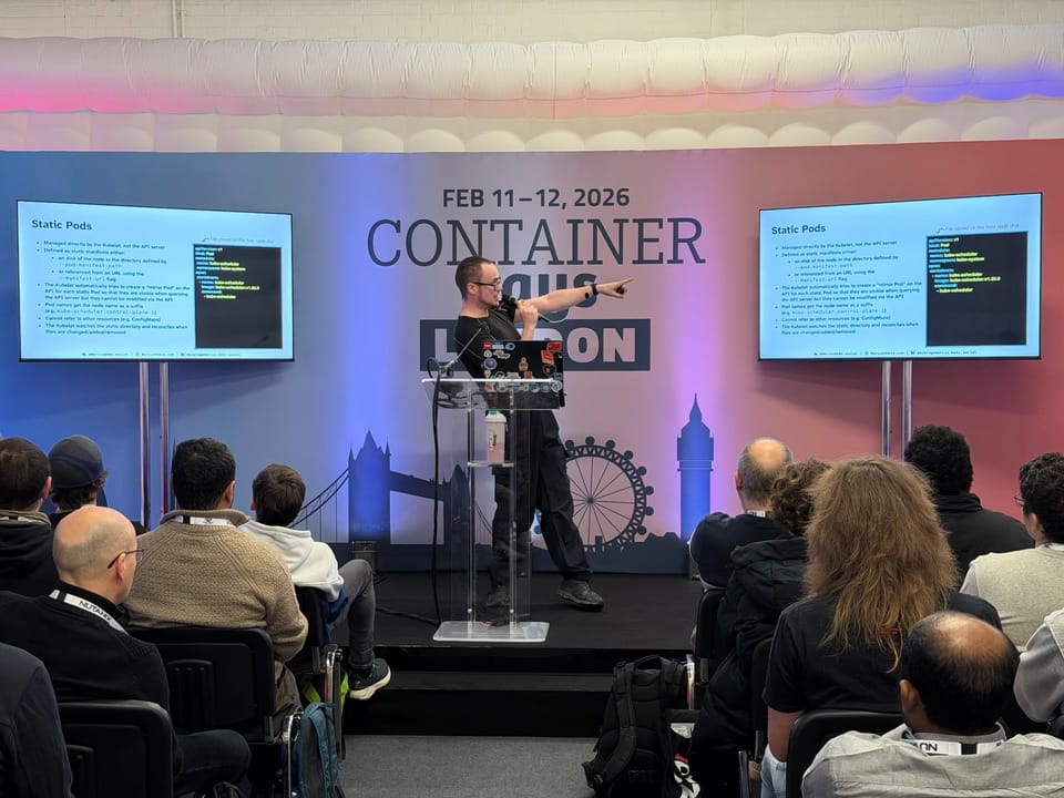 A photo of me stood on a stage behind a podium with two screens positioned at either side of me. I am facing to the side and pointing at one of the screens while holding a microphone.