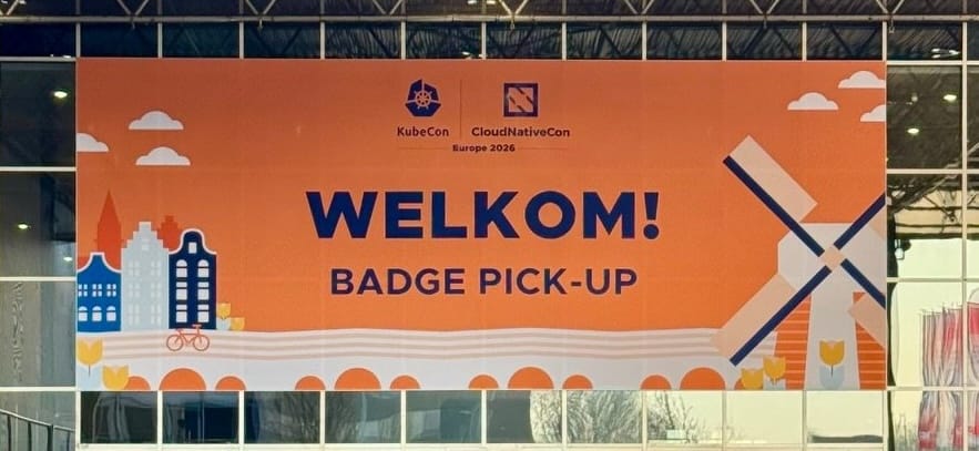 Photo of a large orange banner with the following text in the centre: "Welkom! Badge pick-up". The KubeCon logo can be seen at the top.